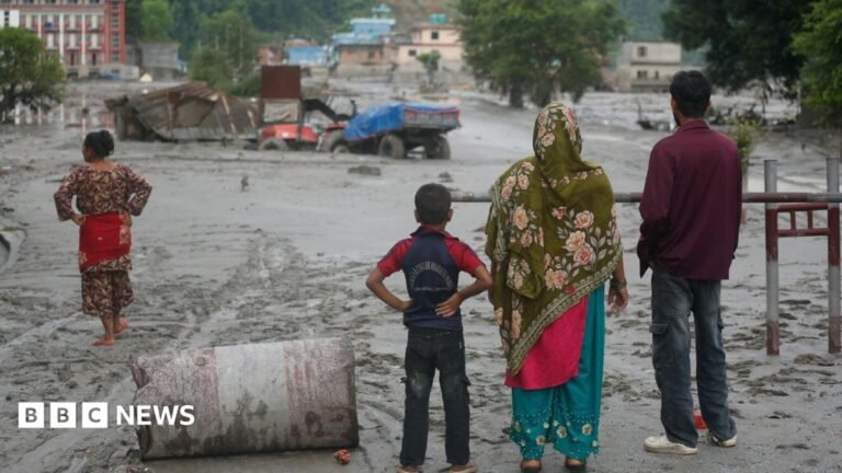 The backs of two women, a man and a boy looking out to partially submerged trucks and buildings as mud covers a section of a highway in Nuwakot in central Nepal