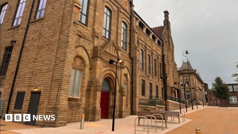 A historic brick building with a red door stands to the left of a public square on an overcast day.