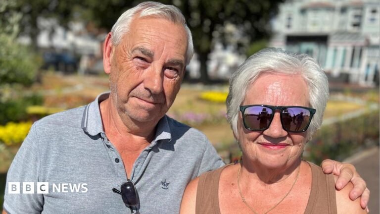 People in Clacton react to Nigel Farage’s by-election win A man and a woman stand behind a trestle table at an election count in a sports hall. They are looking at and holding long ballot papers