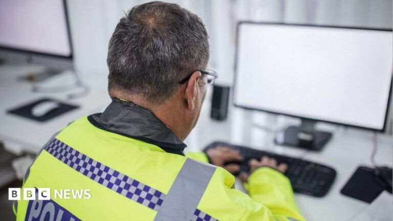 Male police officer wearing high visibility jacket and typing on keyboard as he sits behind computer screen