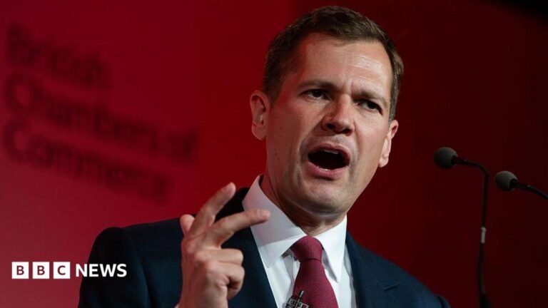 Reform UK benefits ban for foreign nationals would include EU citizens Robert Jenrick makes a hand expression as he speaks at a podium in front of a red-lit background.