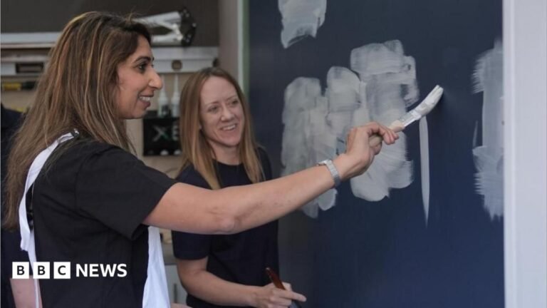 Suella Braverman is wearing a dark-coloured top and white dungarees. She is holding a paintbrush and painting white paint on to a blue wall. She is being observed by a woman in the background, who is wearing a black top.