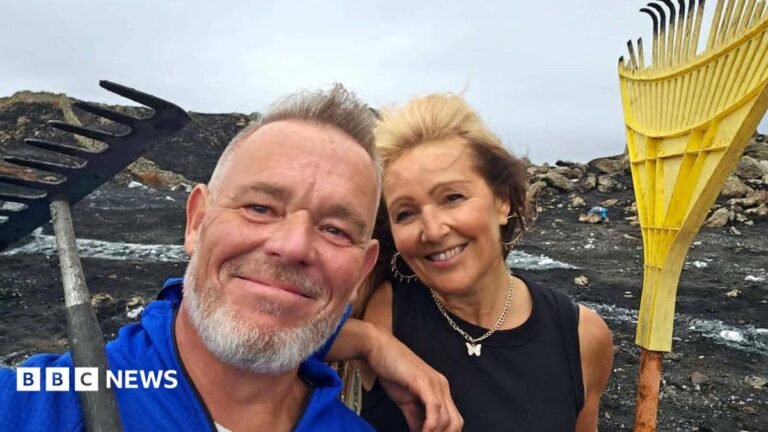 Dean Ward and Allie Evans. He has grey hair and a beard and is wearing a blue top. She has a black top, silver earrings and a silver necklace with butterfly pendant. They are holding rakes and stood on a blackened mountain