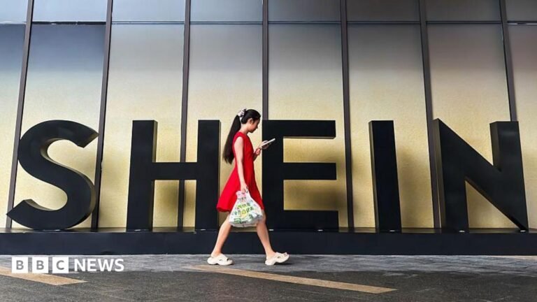 A woman in a red dress looks at her smartphone while walking past a Shein sign outside the company's office in Guangzhou, China.