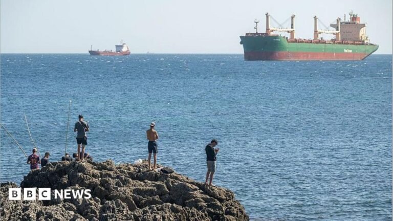 Spanish government says sorry to people of Ceuta for migrant crisis A group of men are seen from the back on a rocky outcrop with the blue sea and large ships in the background, on 22 August, 2026 in Ceuta, Spain.