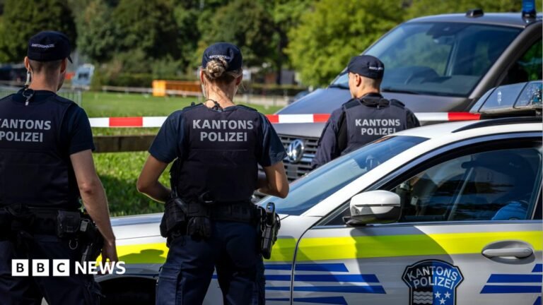 Switzerland rave shooting kills woman, 22 as manhunt launched A close-up photograph shows a man and a woman - both with brown hair - posing together outdoors and looking toward the camera. The man wears a white hat. The backdrop is a city street with a building façade, outdoor seating and an orange scooter visible in the background.