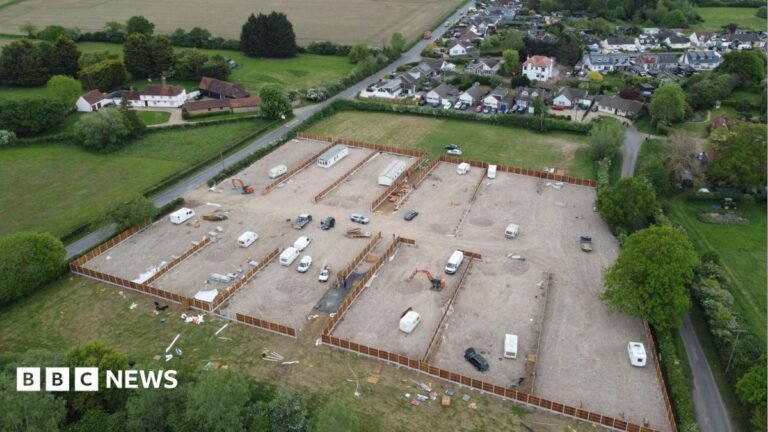 An aerial view of a piece of land that houses several static and touring caravans. There is a road leading into the site and grey hardstanding can be seen. There are also several cars and some smaller buildings.