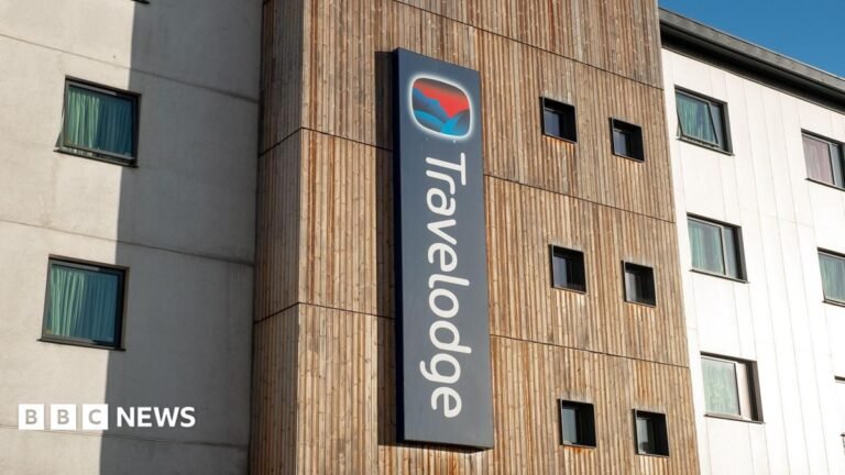 Travelodge boss steps down after security issues in rooms A man in a blue T-shirt, arms crossed, looking serious, standing outside a Travelodge hotel.