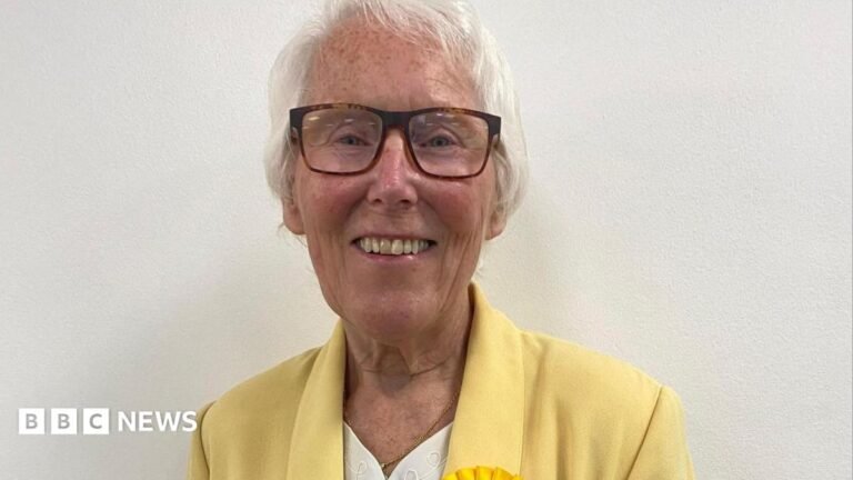 A smiling woman wearing glasses with short, grey hair. She is wearing a cream top underneath a yellow jacket with the top of a yellow rosette showing.
