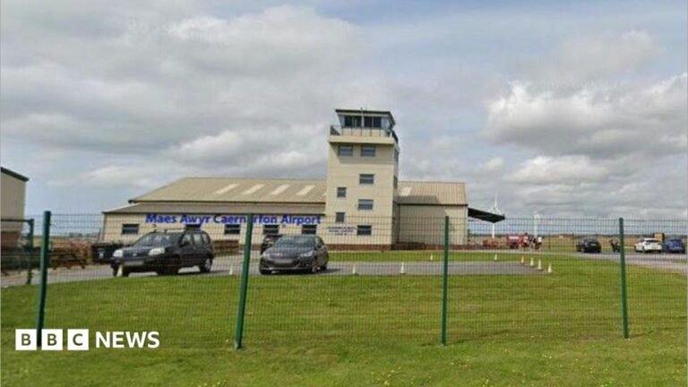 A small cream building airport. It has a watchtower and surrounded by green grass and fields. There's a car park with multiple cars around the building.