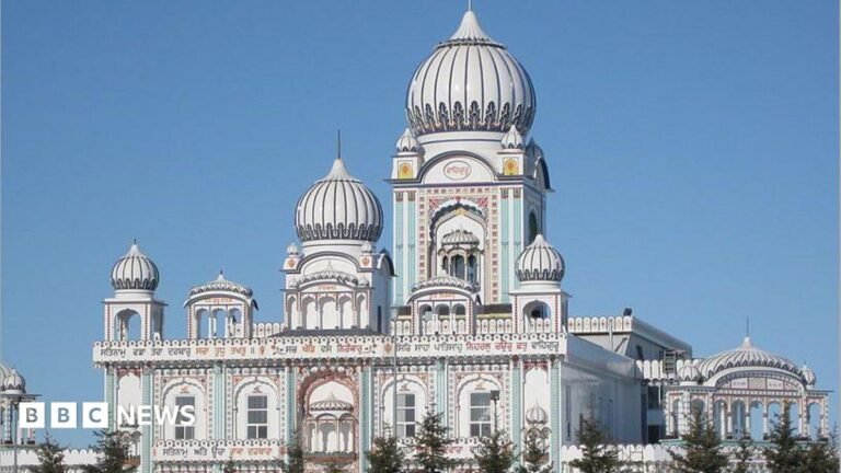 An image showing the outside of the Nanaksar Gurdwara Gursikh Temple Edmonton. It is a large, ornate white building with baby blue and rose colourings on its facade, as well as arches and several domed roofs. It is seen surrounded by spruce trees.