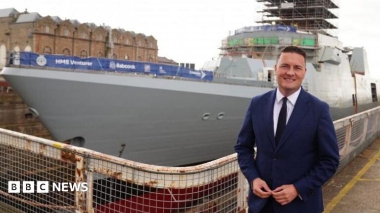 Streeting is smiling and standing in front of a war ship in dry dock, during a visit to a shipbuilding facility in Rosyth. He is wearing a navy suit with a white shirt and a black tie. He has brown, slicked-back hair.