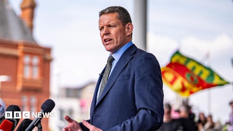 Rhun ap Iorwerth speaking into a set of microphones in front of the Pierhead building in Cardiff Bay, which is out of focus. A group of supporters can be seen behind him, also out of focus.