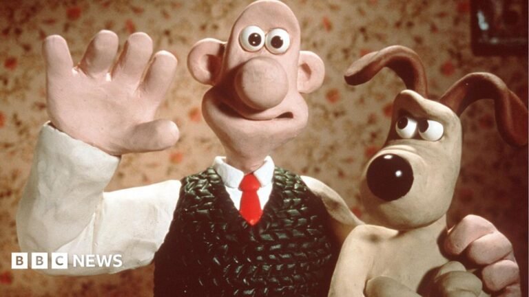 Aardman at 50: Gromit was nearly a cat, Shaun the Sheep almost spoke Mocha wears a fushia pink off-the-shoulder dress with matching pink gloves and has a large brunette wig on. She has dramatic eye makeup. She is standing in front of a poster board which has her face on it.