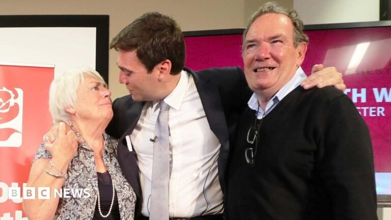 Andy Burnham hugs his parents, Eileen and Roy, after he was selected as the Labour candidate to become the mayor of Greater Manchester in September 2026. They are standing in front of a red banner reading "Labour North West".