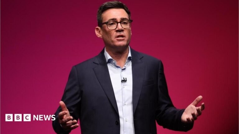 Andy Burnham is wearing a dark-coloured jacket, a white shirt and glasses. He is stood in front of a red backdrop.