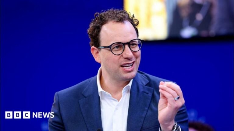 Anthropic boss Dario Amodei calls for AI development to slow down Dario Amodei hld the closed fingers of his left hand together as he mades a point. He is wearing a white shirt and blue jacket, black-rimmed glasses and has slightly curly hair. He is sat against a blue background, with a yellow-lit square just behind and to the left of his head.