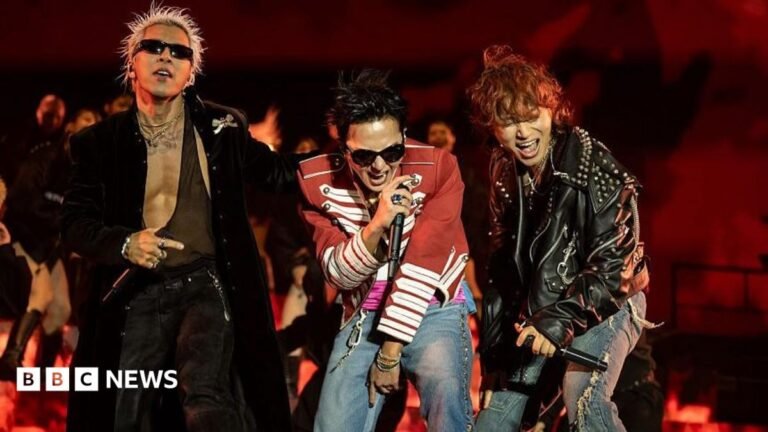 Three performers stand at the front of a stage holding handheld microphones during a live performance. One performer wears a long black coat and sunglasses, another wears a red military-style jacket with white trim and jeans, and a third wears a black studded leather jacket and ripped jeans. The stage is lit in red, with flames or flame-effect visuals displayed behind the performers.