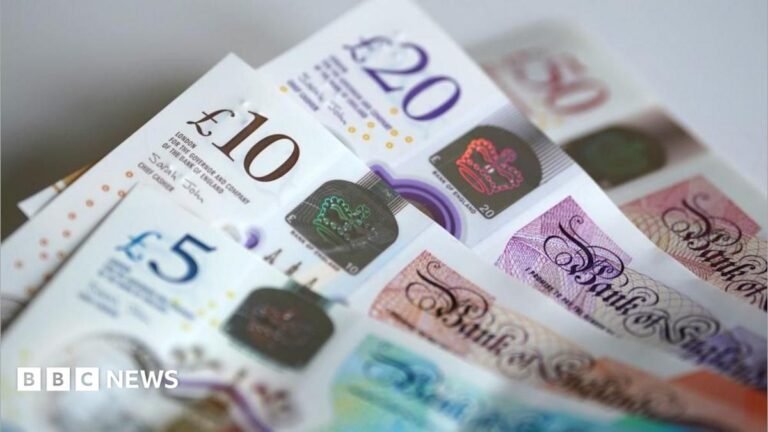Four banknotes are in a pile on a table. Each banknote is partially obscured. There is a £50 note in the background which has a £20 on top of it. A £10 is on top of the £20, and a £5 is on top of the £10.