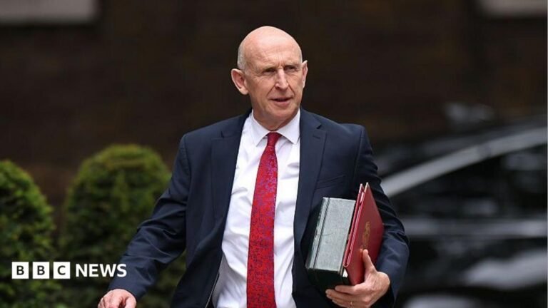 Chancellor John Healey to say UK economy ‘turning a corner’ despite debt concerns An image of John Healey from the waist up. He is walking and wearing a dark suit with a white shirt and red patterned tie. He is carrying a red folder. The background is blurred by you can see that he is outside from the outline of bushes.