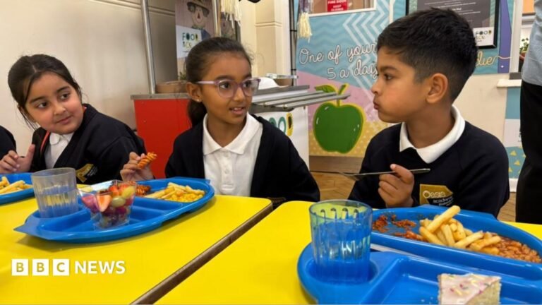 Deep-fried food banned under new school dinner rules in England Three primary school pupils in uniform sit at bright yellow tables in a school canteen, eating lunch from blue trays with chips, fruit and dessert while talking together.