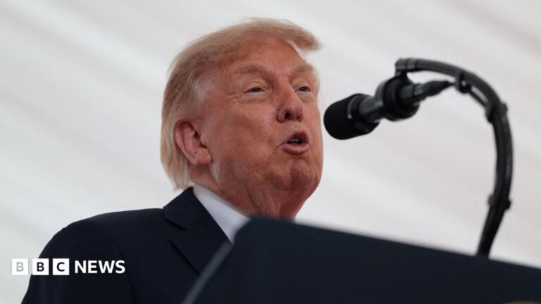 Trump is speaking into a mic. He has a black suit on with a white shirt. A white backdrop is behind him.