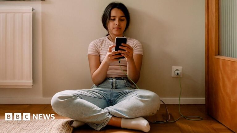 A young girl is seen sat crossed legged on the floor scrolling on a smartphone while it is charging