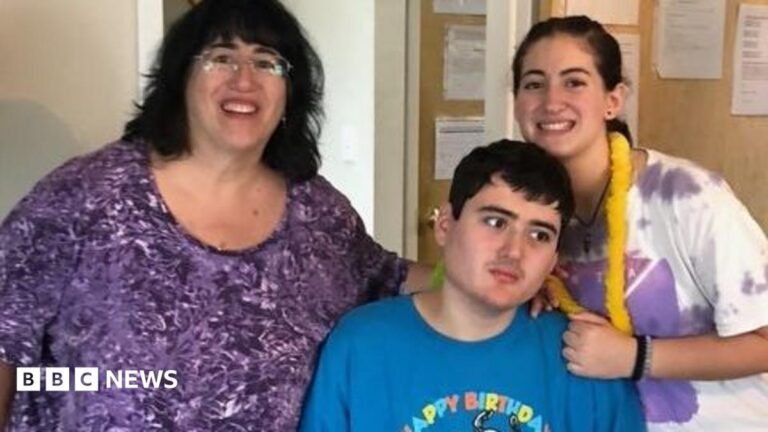 Linda Mancuso, wearing a purple top, glasses and dark hair falling to her shoulders, stands next to her 23-year-old son, Michael, who needs 24/7 care; he wears a "Happy Birthday" blue shirt and stares off to the side; her daughter stands to the other side of Michael, smiling as she rests a hand on his shoulder