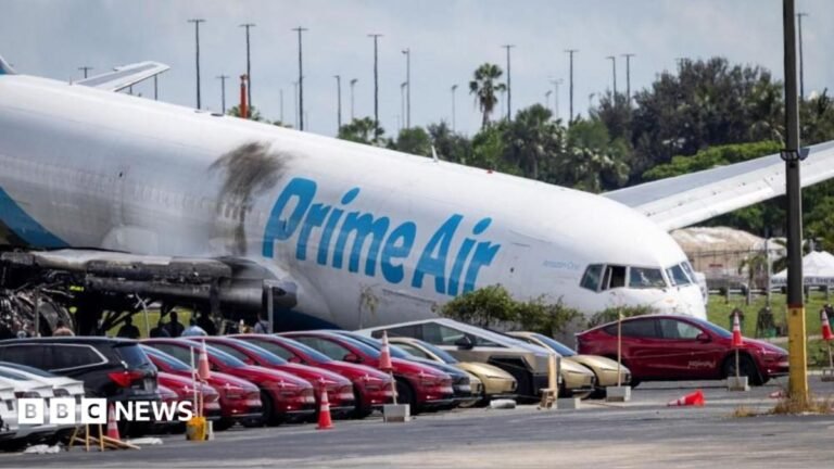 Flight recorders recovered from ‘devastating’ Amazon cargo plane crash A woman with short reddish hair with a neutral expression faces the camera while sitting a room with black and brown wallpaper.