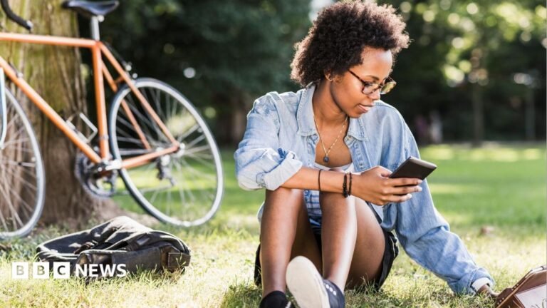 Student sits on the grass with a phone in her hand, a tablet computer on the grass and a bike resting on a tree behind her.