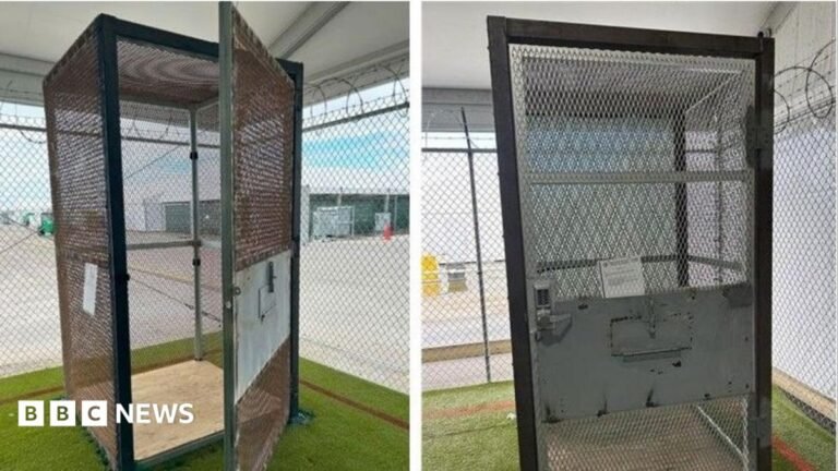 A metal cage sits within a larger cage that has razor wire across the top. The small cage has a door with a combination lock