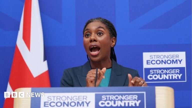 Kemi Badenoch accuses Andy Burnham of kicking defence spending into the long grass Badenoch is pictured from below the podium where she is giving a speech, at London Scottish House in Westminster, with a royal blue background and a Union flag to her left. She wears her braided hair tied back in a low ponytail, with a navy jacket and a turquoise top.