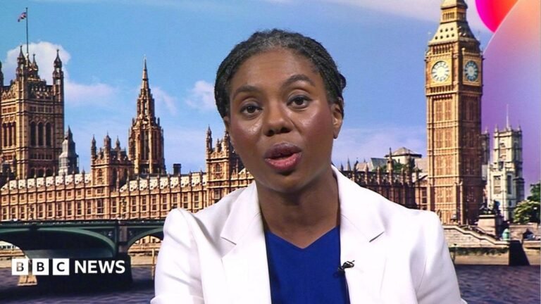 A head and shoulders shot of Badenoch in a BBC studio. She is wearing a white jacket over a royal blue top and has her braided hair tied back in a low ponytail.