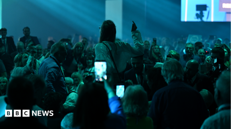 Man cautioned over Reform conference assaults in Birmingham A woman stands in a crowd in a darkened hall and appears to be pointing and shouting. Several people are standing around her, some with phones held up to film.