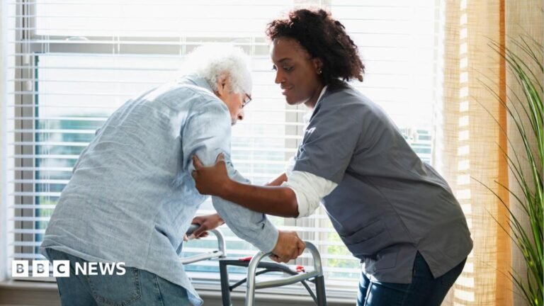 A female care home worker, wearing nursing uniform, helps support an elderly man leaning on a walking frame.