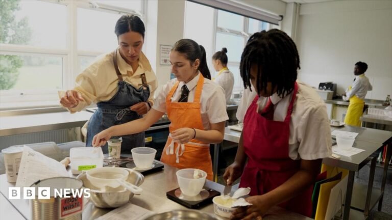 Two teenagers at a classroom bench, wearing aprons., They are in a catering class and are adding cream cheese to bowls, and are being supervised by their female teacher, also in an apron, who is standing to the left of them.