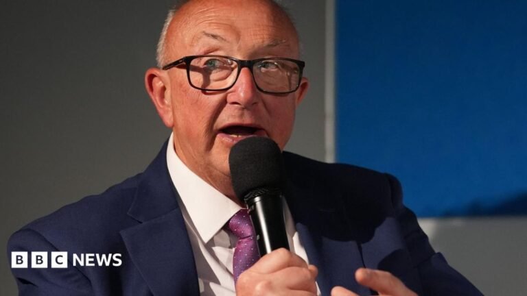 Norfolk police commissioner cancels ‘ridiculous’ theatre tour Colin Sutton is wearing black rimmed glasses and a navy suit, white shirt and purple patterned tie. He is holding a microphone close to his mouth and looks to be in a corporate environment.