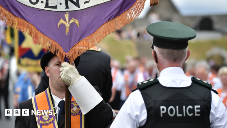 Orange Orderparade along Garvaghy Road halted after injuction granted A man is wearing an orange sash and a black bowler hat and holding a banner. A policeman has his back to the camera. He has a black vest on stating police in white lettering. He has a cap and a white shirt on. A parade is out of focus in the background.