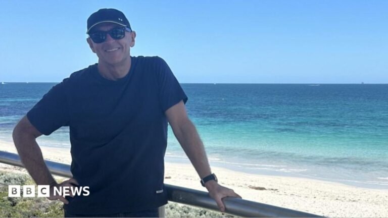 Greg O'Neill stands beside a beachfront railing overlooking clear blue water and white sand on the Western Australian coast.