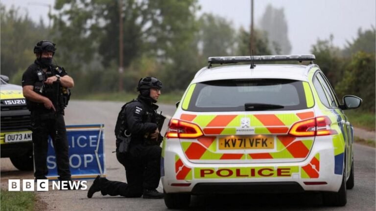RAF Fairford: ‘Quantity of petrol’ but no explosives found in three vehicles An armed officer stands in uniform as another one crouches down with a gun in his hand pointed down and he talks through the window of a police car which has its back with a sign 'police' visible