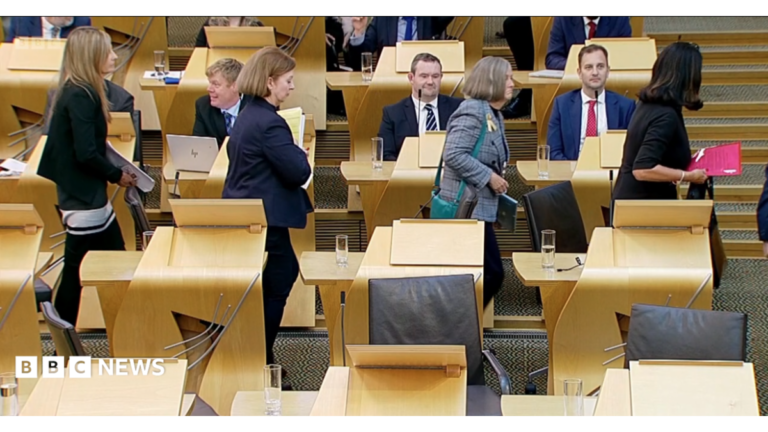 John Swinney, who is bald with glasses and wearing a navy business suit with purple tie, stands in a debating chamber.