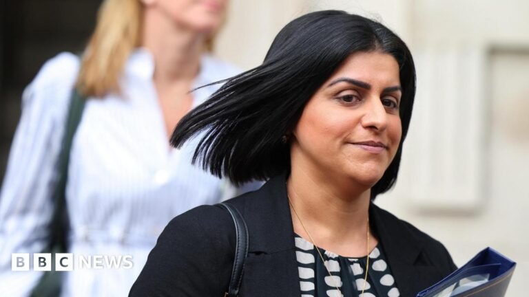 Shabana Mahmood to reopen scheme to resettle refugees in the UK Shabana Mahmood is holding a blue folder. She is wearing a black jacket, a black and white top, and has a bag strap over her right shoulder.