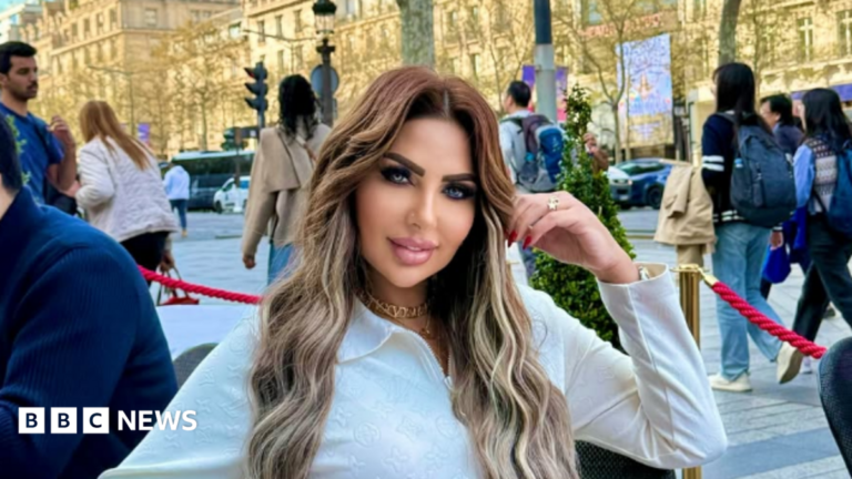 Sarah Khalifa wearing a white long-shirted with her folded left arm, the hand touching her long hair, sits by the side of a road inside a red-roped cordoned area, beyong which people walk on a road the other side of which has rows of sunbathed buildings