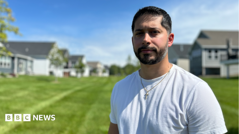Gerald Bourguet, a white man standing in a field of a manicured lawn. He is wearing a white shirt and crucifix chain. There are homes in the background that are blurred.
