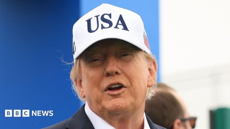 U.S. President Donald Trump attends the Amgen Irish Open at Trump International Golf Links in Doonbeg, Ireland. He is wearing a white USA baseball cap, navy blazer with a white shirt. He has his arms outstretched.