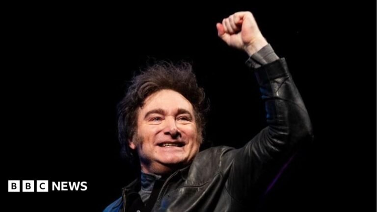Argentine President Javier Milei, wearing a leather jacket, stands smiling with a closed fist raised against a black backdrop.