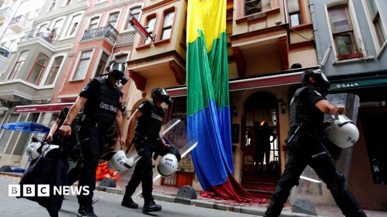Turkish police detain dozens in raids on gay bars and homes of LGBTQ+ activists Riot police, with a rainbow flag in the background, chase LGBT activists as they try to gather for a pride parade, which was banned by the governorship in Istanbul. Officers are wearing black uniforms, wearing gas masks, and are carrying helmets and riot shields as they move through what appears to be a residential street.