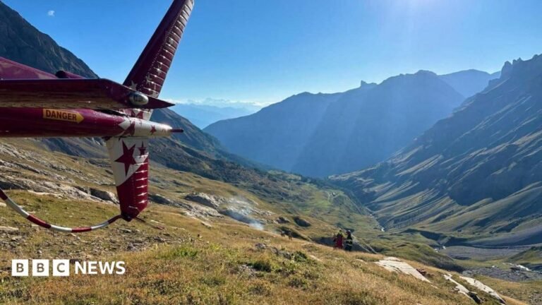 Helicopter tail in foreground with rising smoke in the distance and emergency crews seen high in the Alps overlooking a mountain range