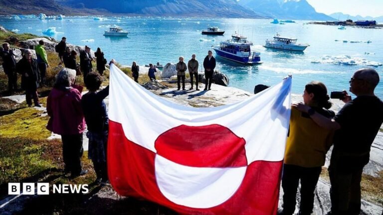 US and Denmark reach deal over Greenland’s security after Trump threats People hold a Greenland flag on the shoreline of Bear Island with boats floating in the background on a sea spotted with large chunks of ice