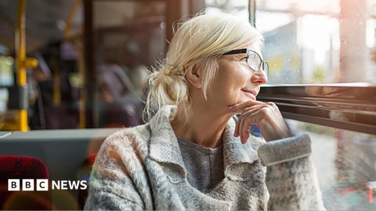 What is the triple lock and why are people talking about it? An older woman smiles as she looks out of a bus window. She has short grey in a bun and is wearing a grey jumper.